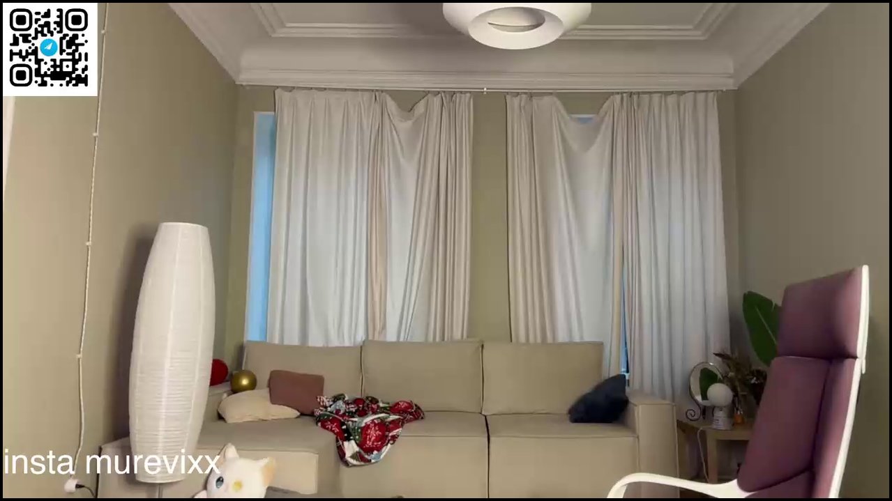 Video of a thin, topless transgender woman with wet hair, wearing black shorts, standing in a dimly lit living room with beige walls, white curtains, 