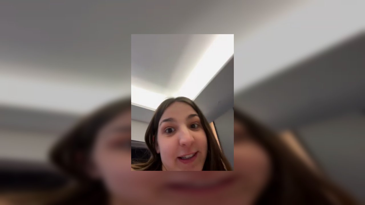 Video of a fair-skinned, topless transgender woman with long brown hair, sitting on a bed with a red floral sheet. She holds a flaccid penis in her ri