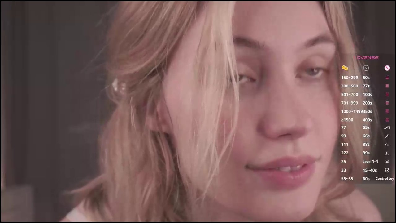 A nude, fair-skinned transgender woman with small breasts and long, blonde hair lies on a bed, masturbating with a black dildo. The background is a be