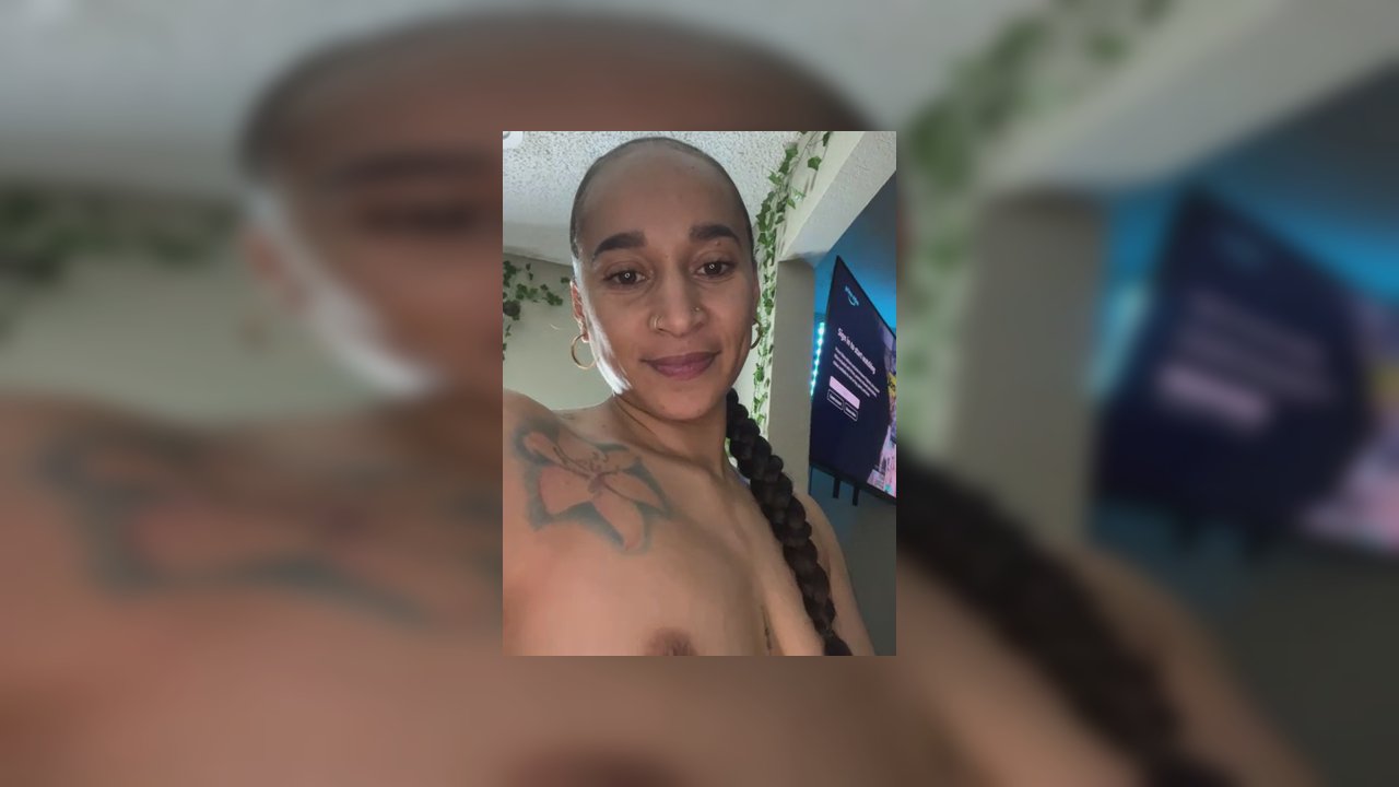 Video of a nude, light-skinned transgender woman with tattoos, lying on a bed, spreading her legs, revealing her vulva, wearing a pink ring on her fin