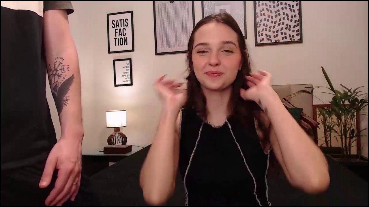 A video of a transgender woman with long brown hair, wearing a black sleeveless top, sitting on a bed in a bedroom. A man's uncircumcised penis is vis