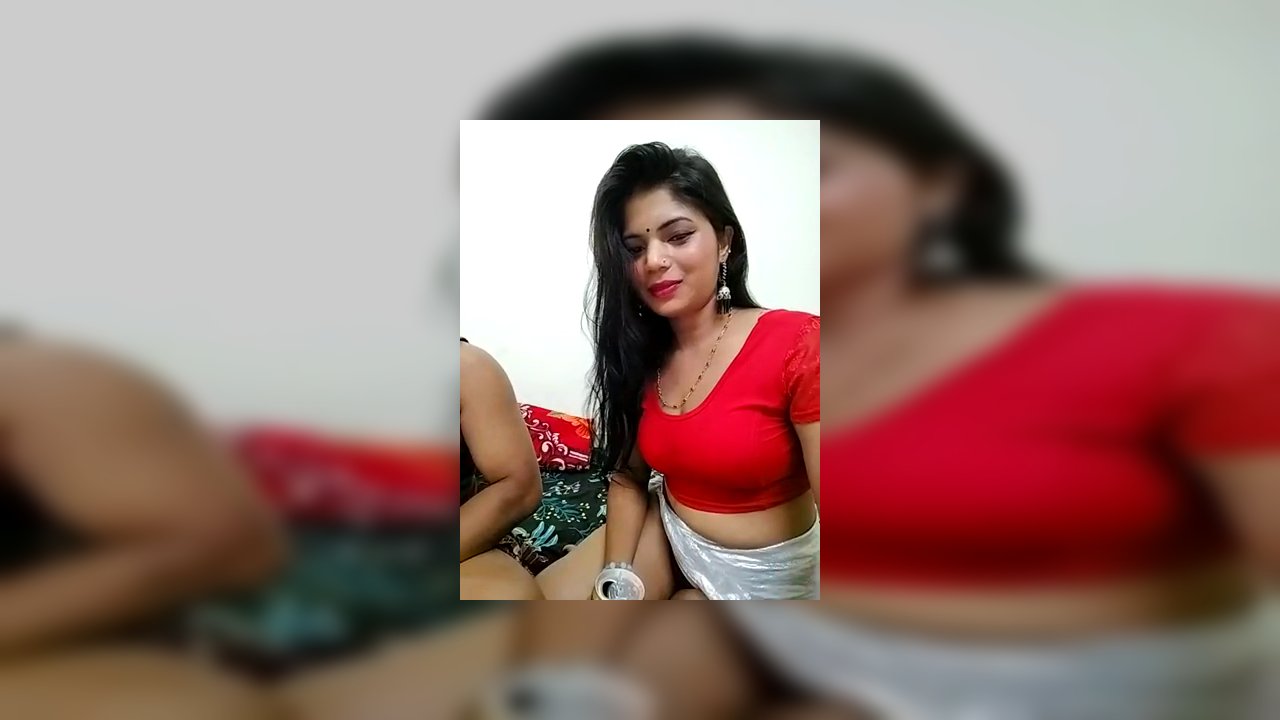 A video of a transgender woman with long black hair, wearing a red crop top and white skirt, sitting on a bed, holding a beer. A man with a dark-skinn