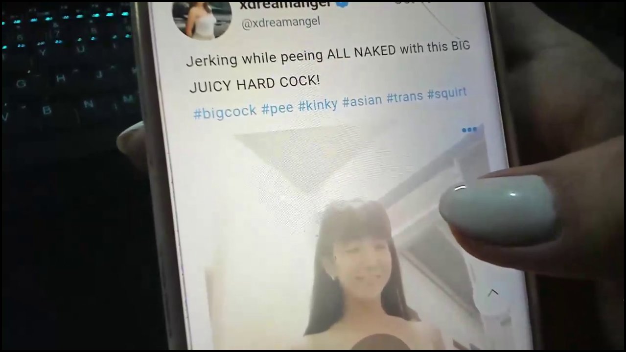 A video of a nude, light-skinned transgender woman with long brown hair, wearing glasses, masturbating with a large, erect penis. She's lying on a bed