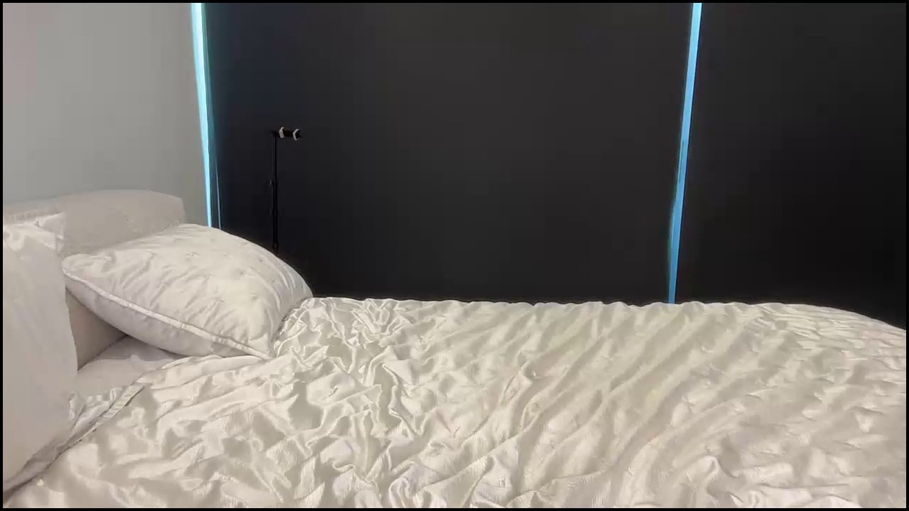 Video of a slender, light-skinned transgender woman with long brown hair, wearing a bright pink bikini, sitting on a bed with white sheets in a modern