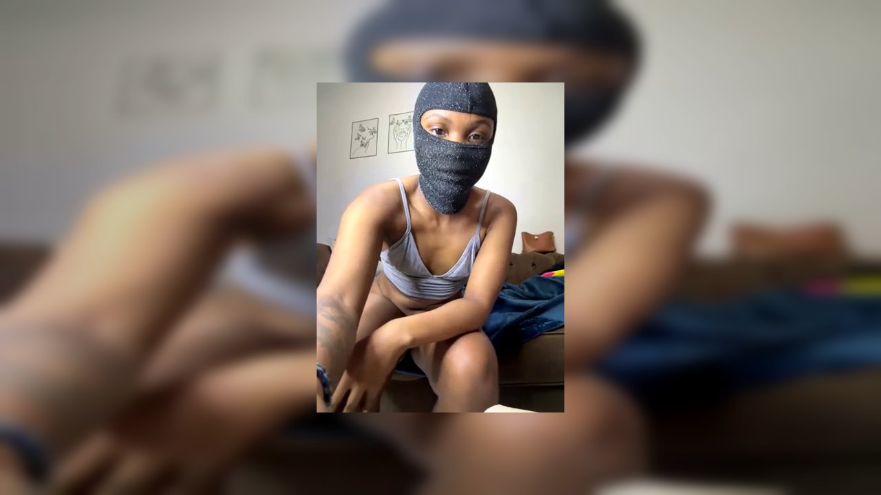 Video of a slender, light-skinned transgender woman with a gray headscarf, wearing a gray bra and panties, adjusting her pants. Background features a 