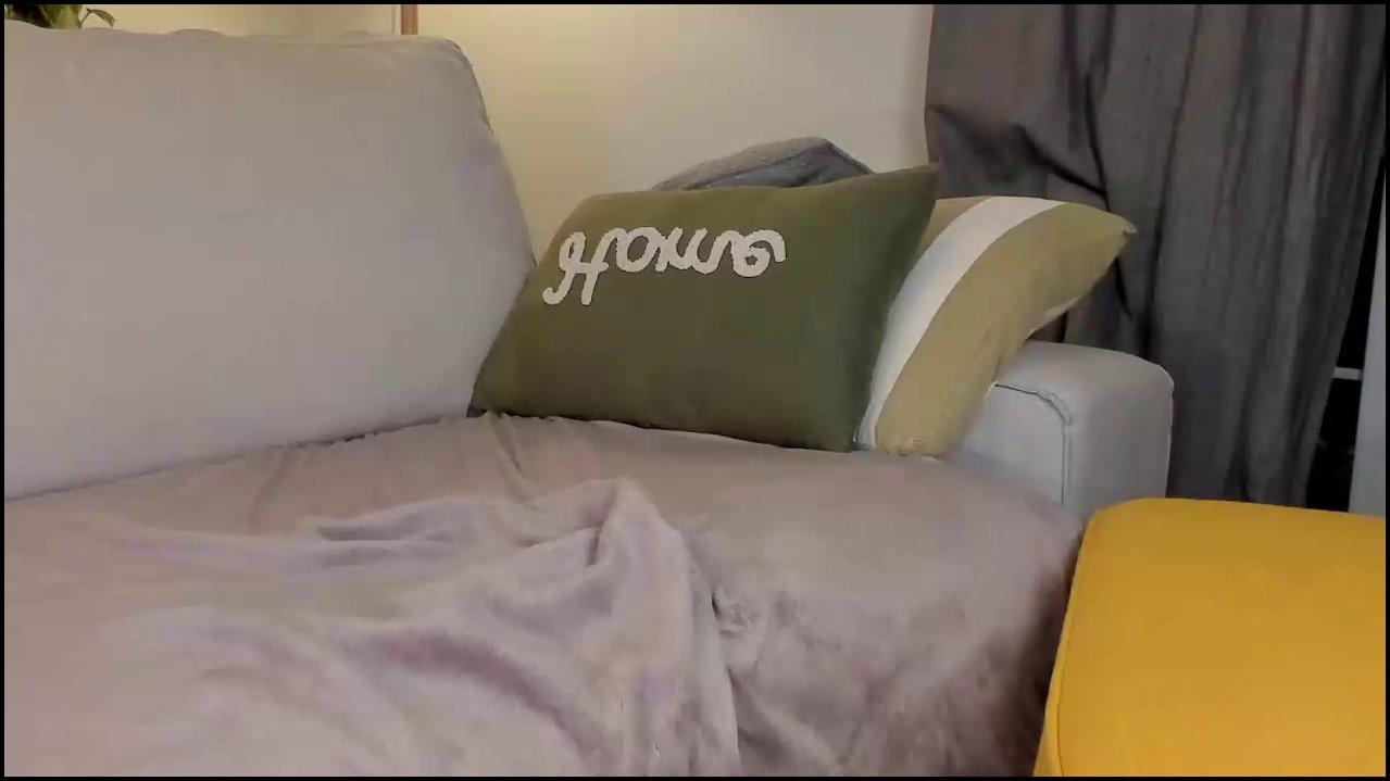 Video of a nude transgender woman with light skin, long brown hair, tattoos, and erect penis, lying on a gray couch, smiling.