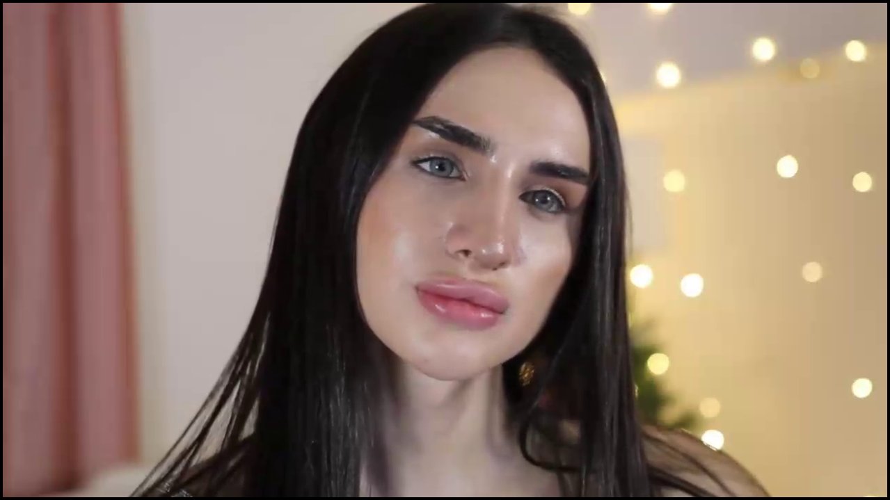 Video of a fair-skinned transgender woman with long, straight black hair, wearing a black shirt, and a subtle makeup look with soft pink lips. Backgro
