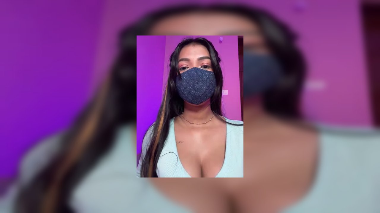 Video of a topless transgender woman with medium brown skin and long dark hair, wearing a blue mask, holding an erect penis, against a gradient purple