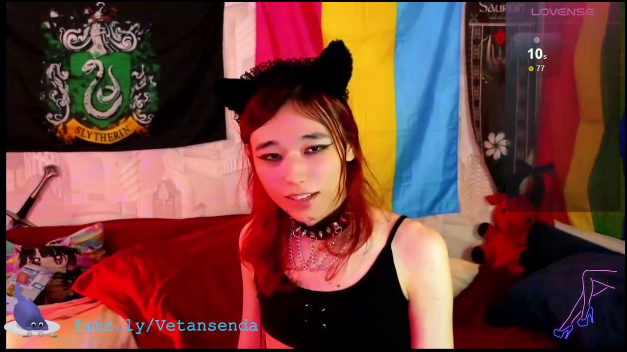 Video of a slender, light-skinned transgender woman with shoulder-length brown hair, wearing a black bra, fishnet stockings, and a collar, masturbatin