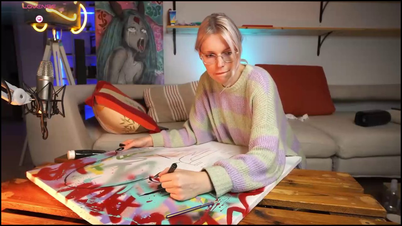Video of a nude, fair-skinned transgender woman with glasses, light brown hair, and a tattoo, sitting on a red chair, legs spread, using a dildo.
