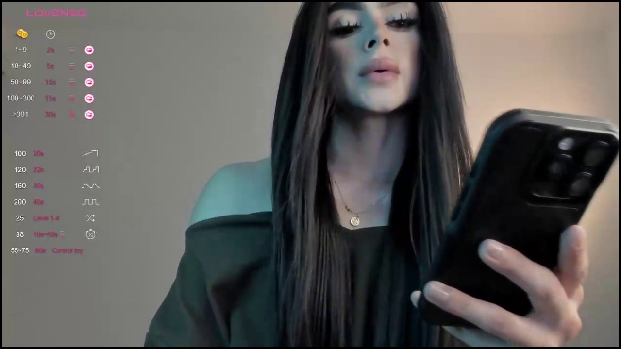 A video of a light-skinned transgender woman with long, dark hair, wearing a green off-shoulder top, sitting on a bed, exposing her erect penis.