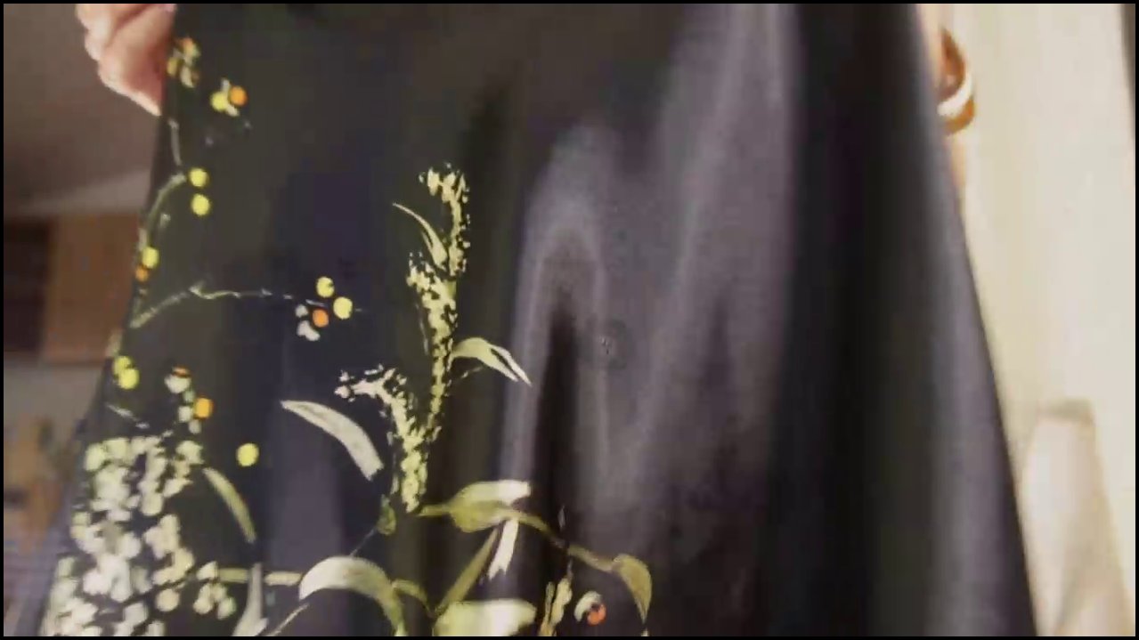 Video of a transgender woman with long dark hair, wearing a black satin top, leopard-print stockings, and a necklace, seated on a white chair, reveali