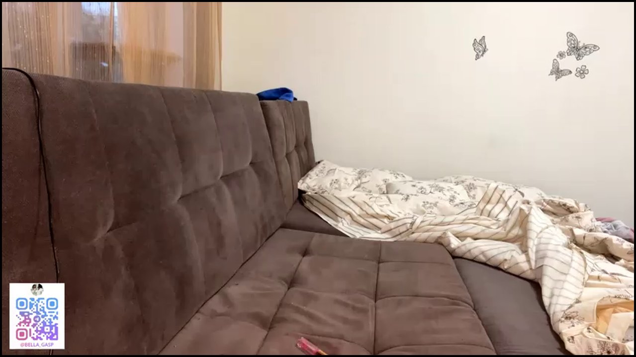 Video of a nude woman with light skin, lying face down on a dark gray couch, legs spread, revealing her anus and vulva. The room has a disheveled bed 