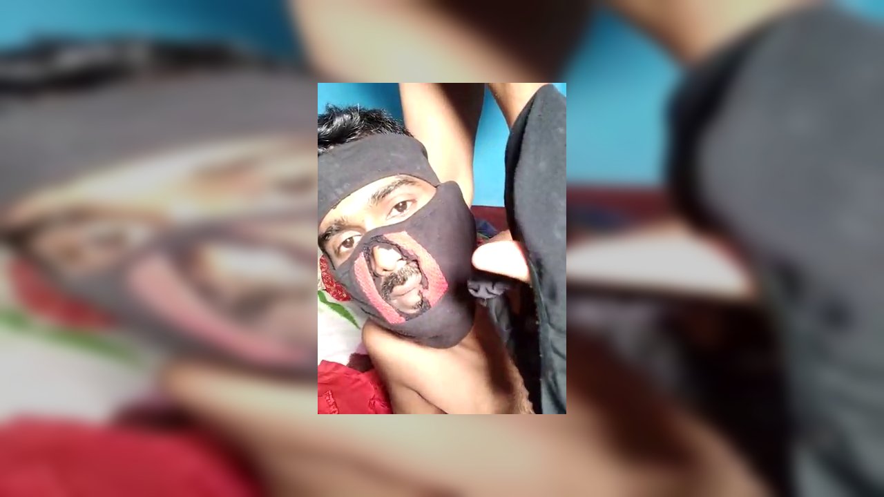 Video of a person, possibly transgender, wearing a red and white patterned headscarf, blue nails, and a black hat, sitting cross-legged on a bed with 