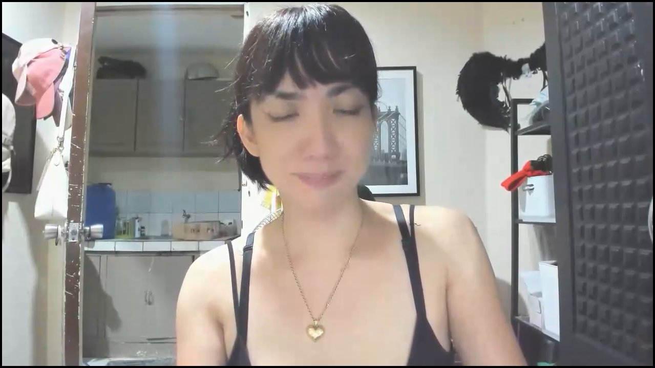 Video of an Asian transgender woman with long black hair, small breasts, and erect penis, lying naked on a bed with white and black sheets, smiling. B