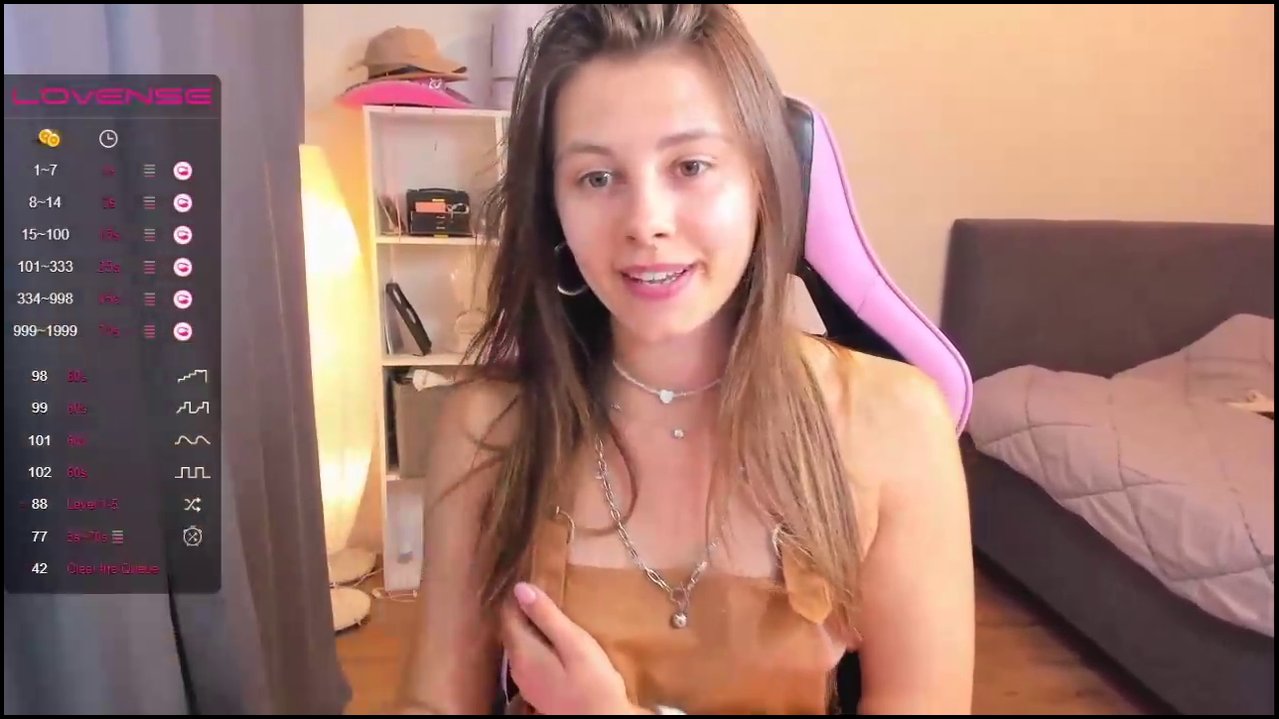 A video of a nude transgender woman with long brown hair, sitting on a pink gaming chair, pulling down white panties, revealing a penis.