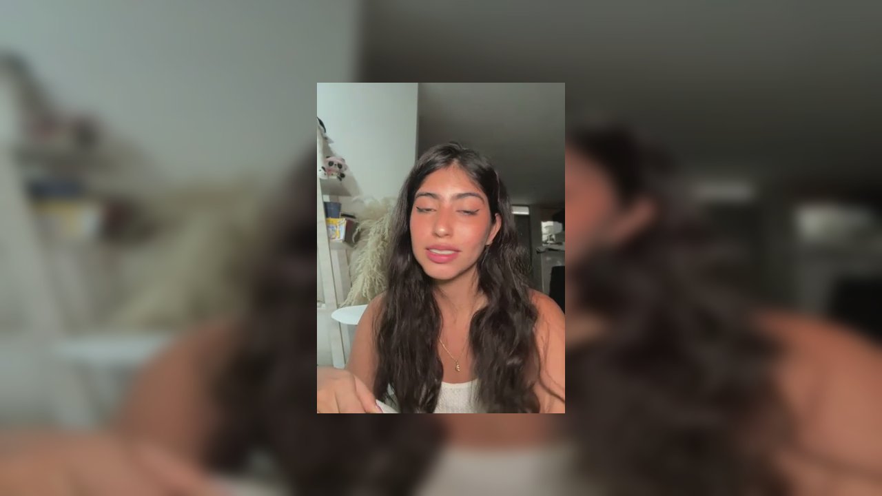 Video of a nude, light-skinned transgender woman with long, curly hair, seated on a bed, legs spread, penis visible, in a dimly lit bedroom with scatt